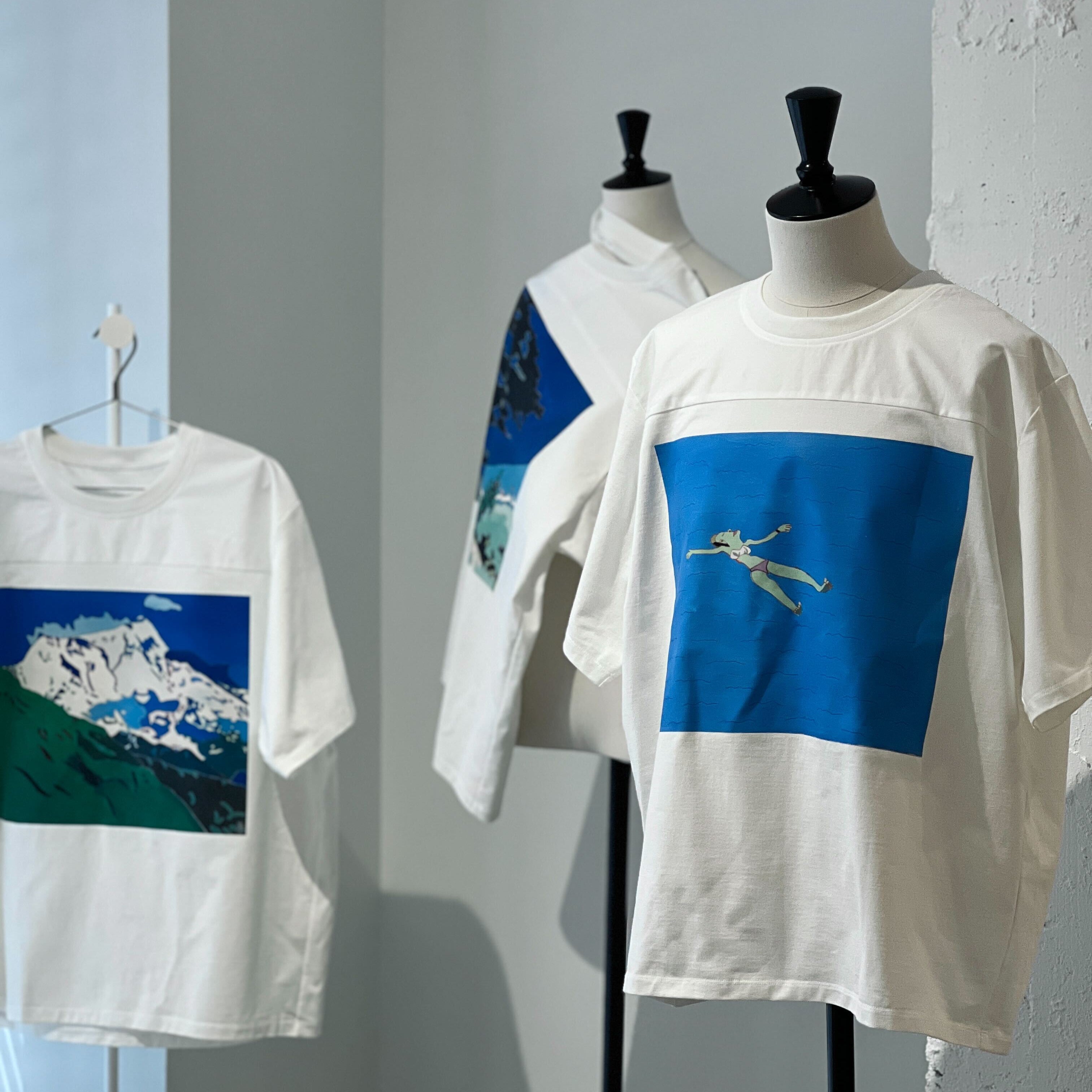 画像:ヘラルボニー銀座店に並ぶ、限定Tシャツ三枚。それぞれ、自閉症の作家こばやしやすひろによる青いアートが正面にプリントされている。画像おわり。